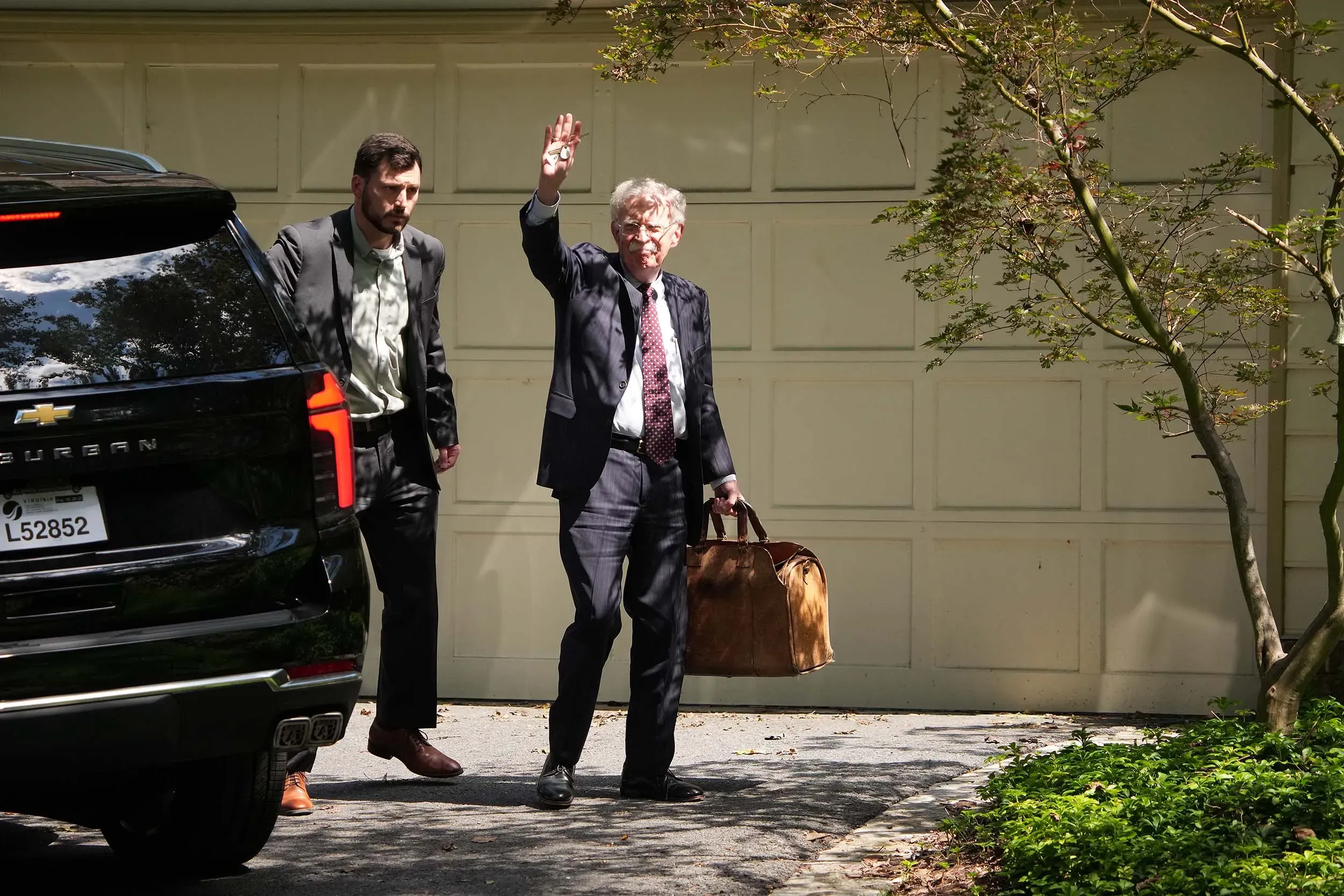 جون بولتون يغادر منزله في بيثيسدا بولاية ماريلاند، حاملاً حقيبة، بينما يلوح بيده، وسط تحقيقات فدرالية حول الوثائق السرية.