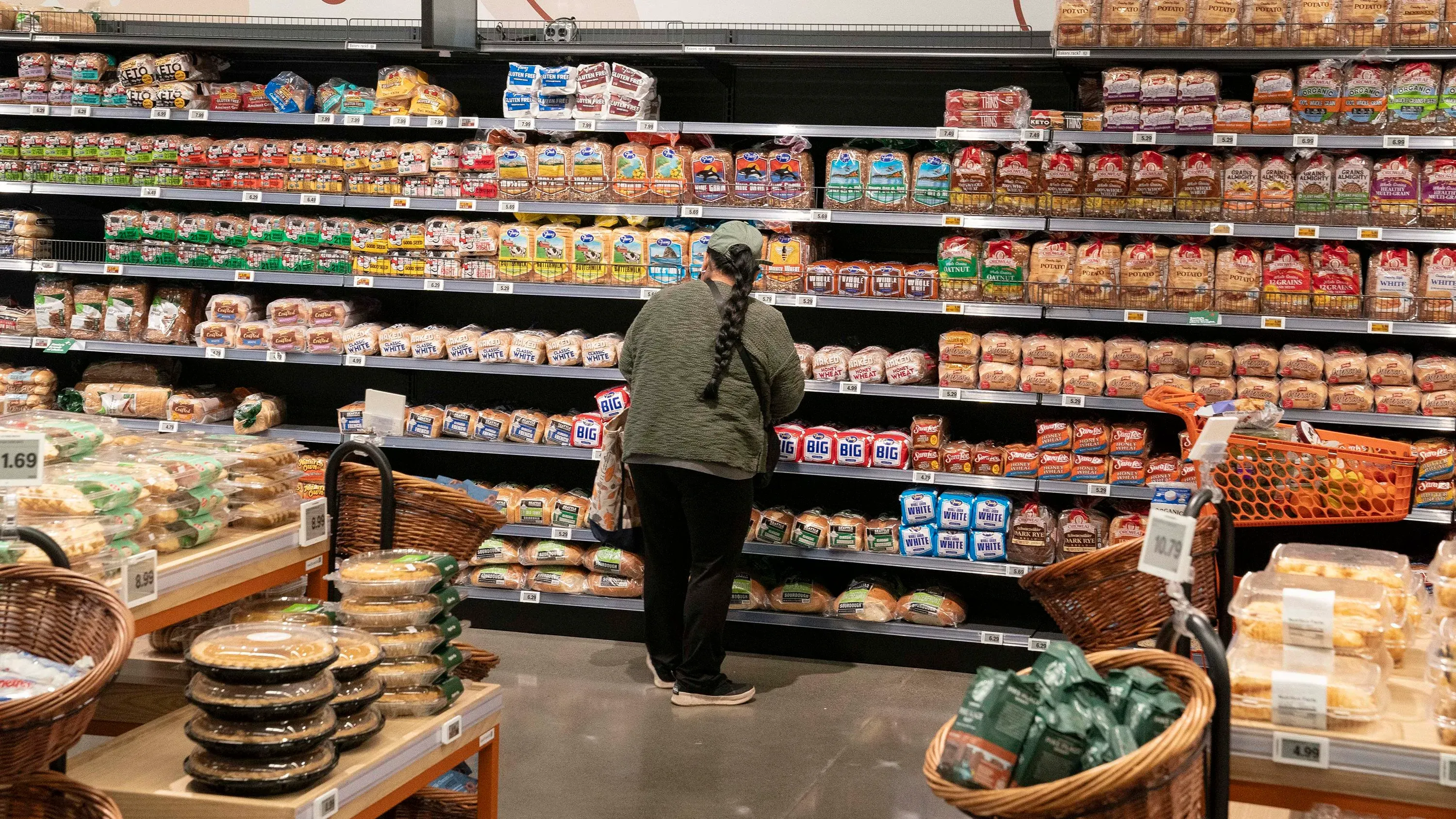 امرأة تتسوق في متجر، تتفحص رفوف الخبز المعبأ، مع وجود سلال مليئة بالأطعمة في المقدمة، في سياق تحديث معايير التغذية.