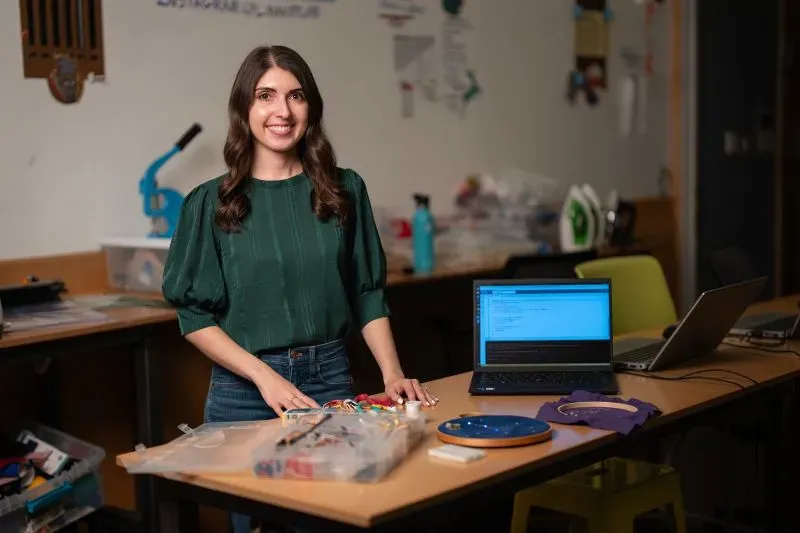 امرأة شابة مبتسمة ترتدي قميصًا أخضر، تقف أمام طاولة مليئة بالأدوات الإلكترونية، مع جهاز كمبيوتر محمول يعرض برمجة، في مختبر تصميم.