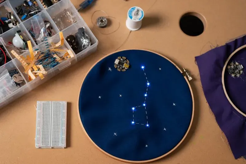 مشروع تصميم أزياء يجمع بين الخياطة والتكنولوجيا، يظهر قماشًا أزرق مزينًا بأضواء LED تمثل كوكبة، مع أدوات كهربائية بجانبه.