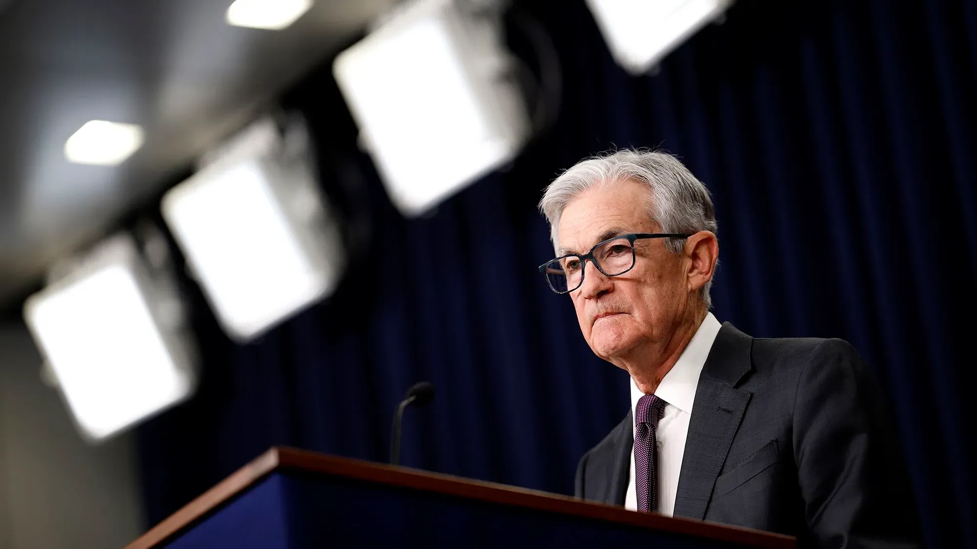 جيروم باول، رئيس مجلس الاحتياطي الفيدرالي، يتحدث خلال مؤتمر صحفي، مع أضواء ساطعة وخلفية زرقاء، معبرًا عن تحديات السياسة النقدية.