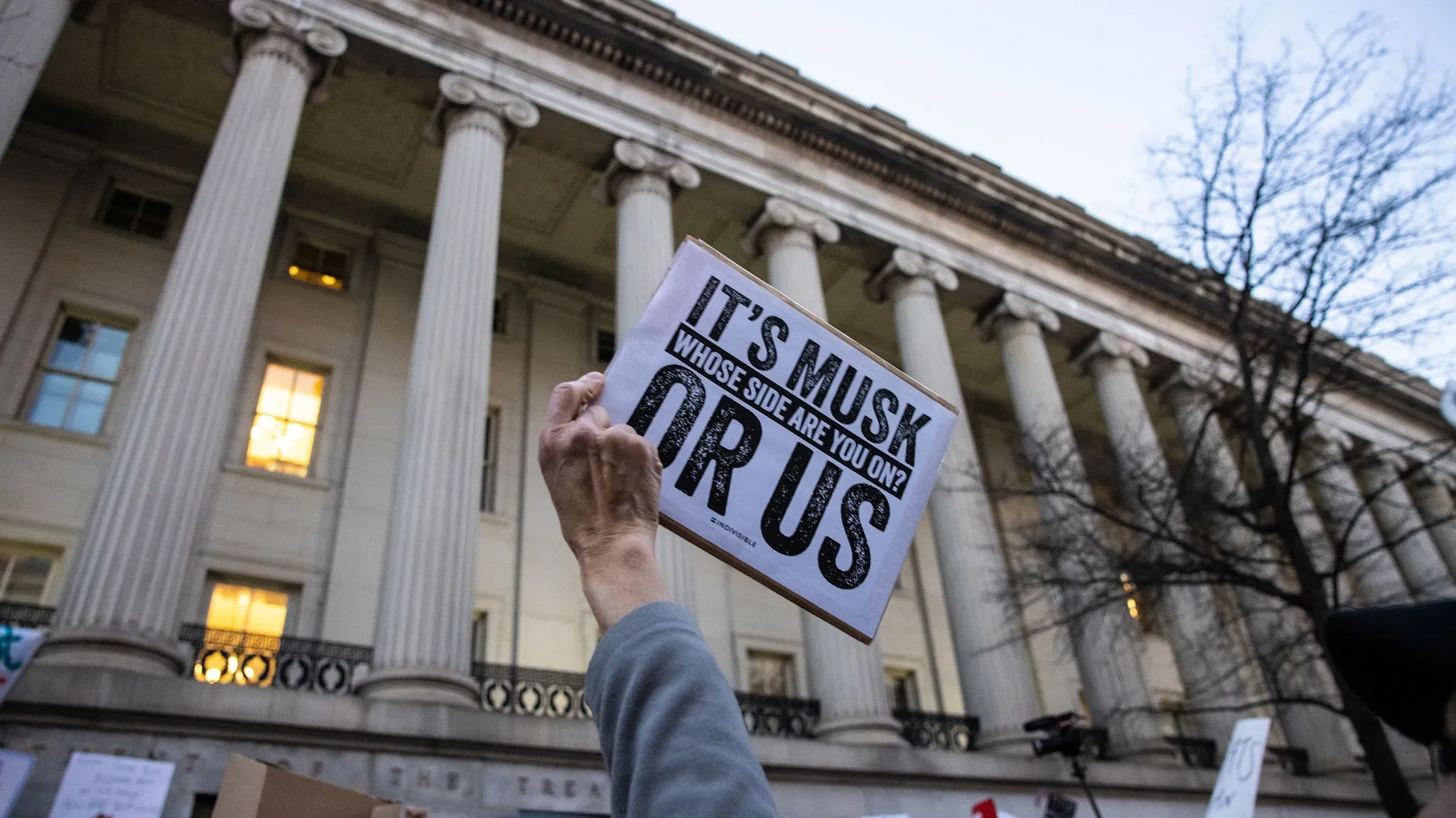 لافتة تحمل عبارة "إنه ماسك، أي جانب أنت؟" أمام مبنى وزارة الخزانة، تعكس الجدل حول وصول فريق ماسك إلى نظام الدفع الحكومي.