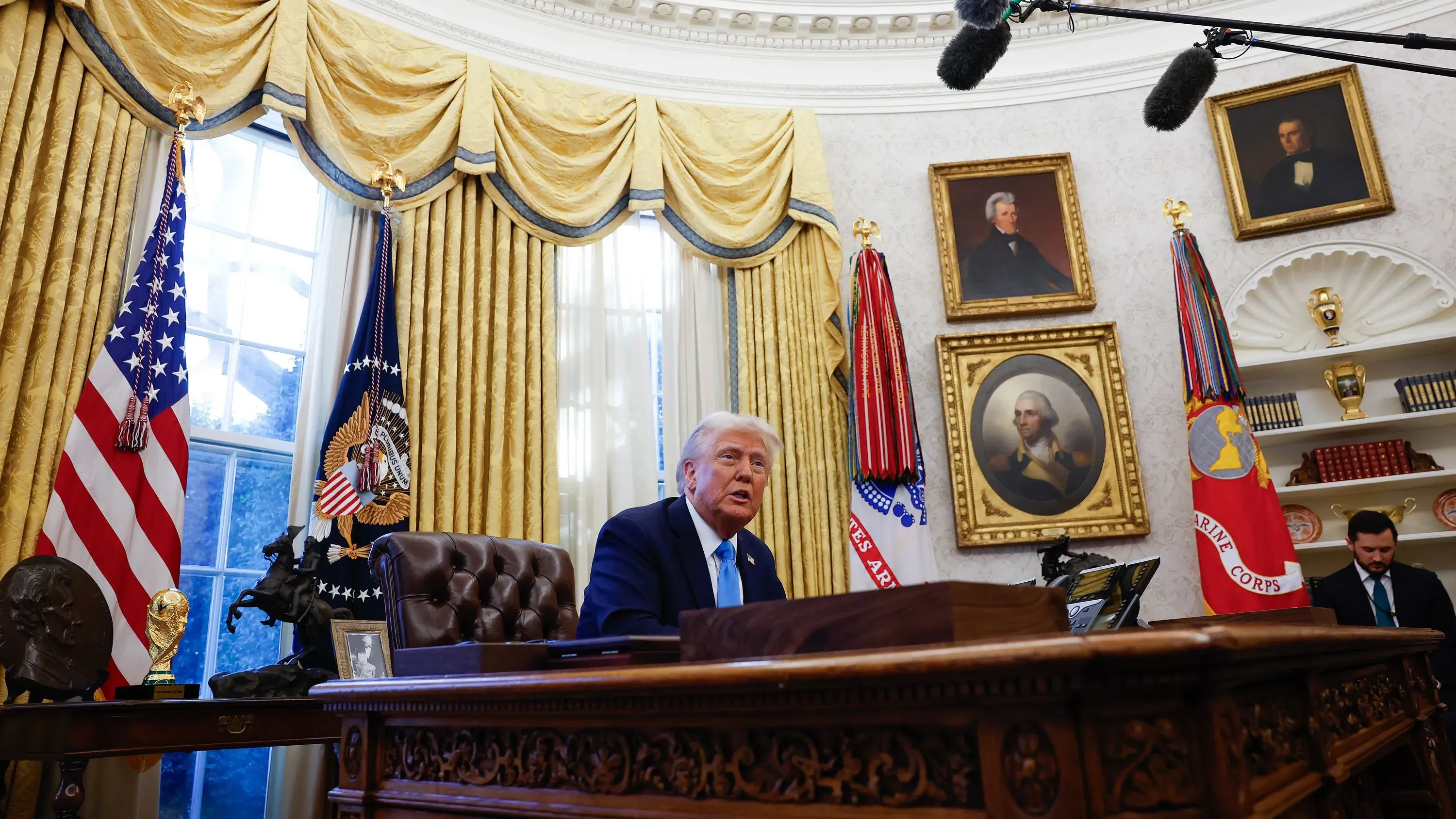الرئيس دونالد ترامب يجلس خلف مكتبه في المكتب البيضاوي، محاطًا بالستائر الذهبية، مع العلم الأمريكي وشعار الرئاسة خلفه.
