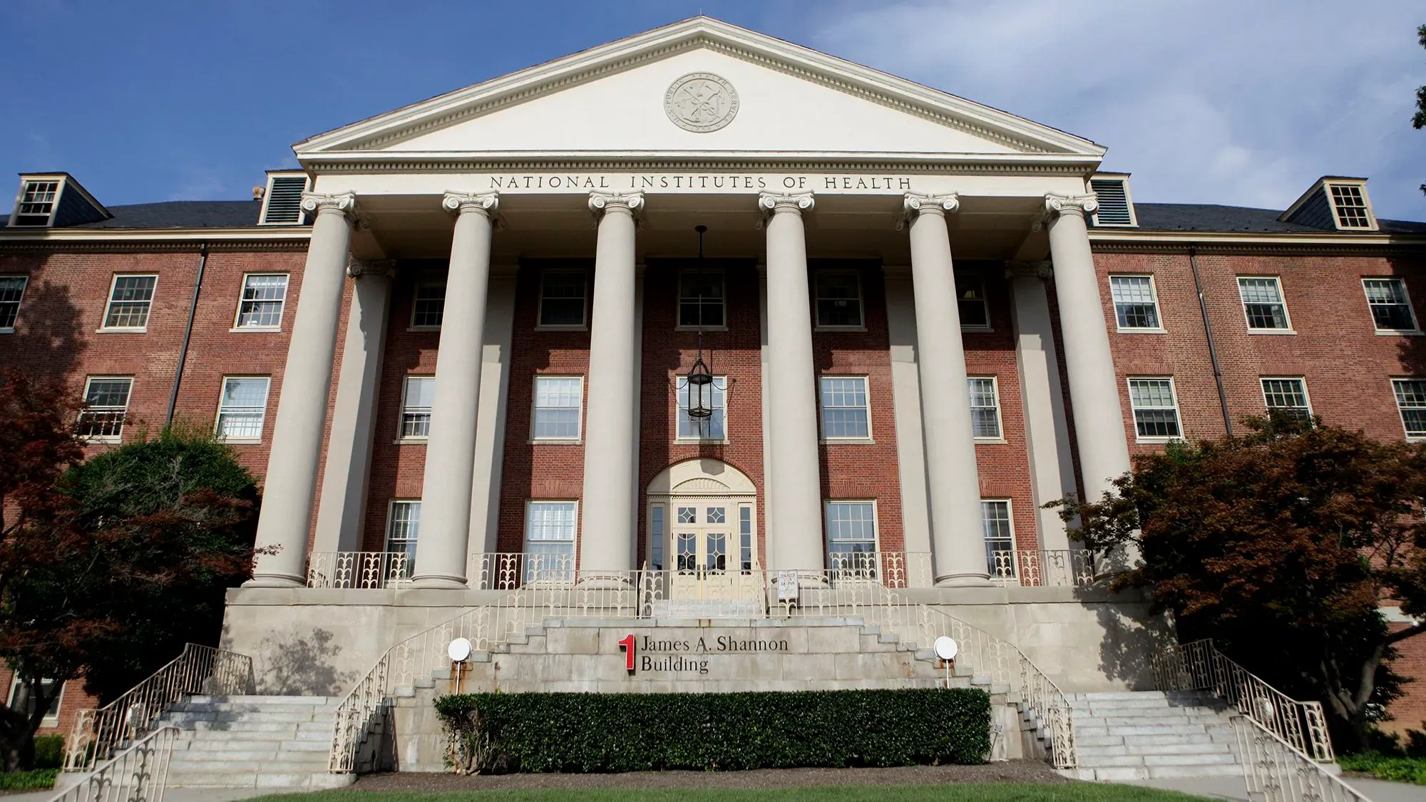 مبنى المعاهد الوطنية للصحة يظهر بوضوح، مع أعمدة بارزة وشعار المعهد، في سياق الجدل حول تمويل الأبحاث الطبية في الولايات المتحدة.