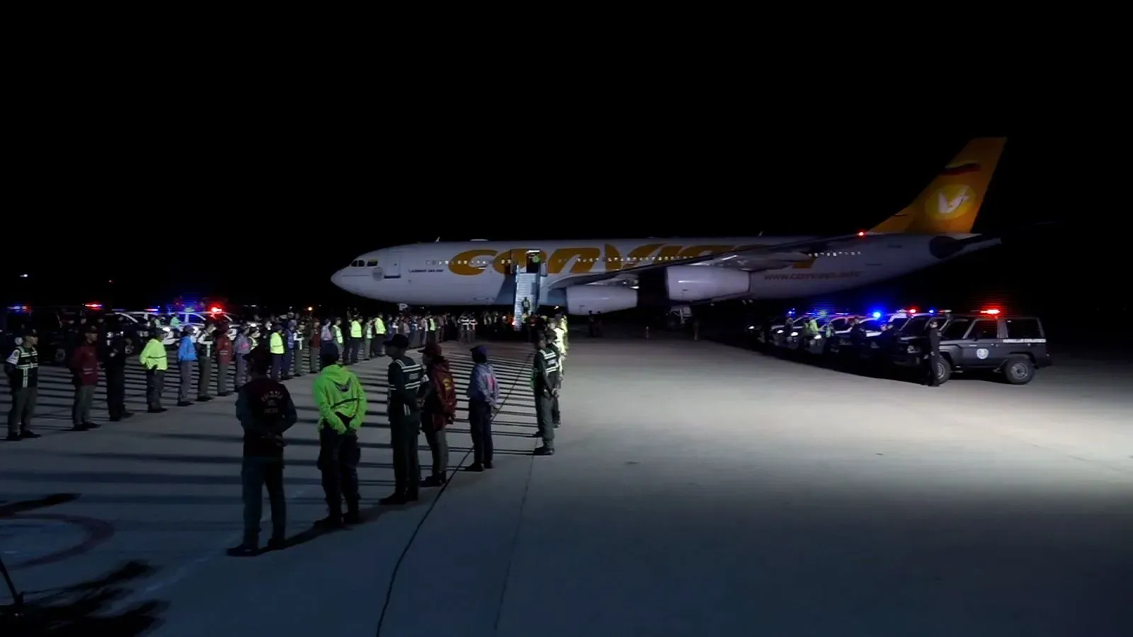 مطار ليلي مزدحم، حيث تقف قوات الأمن بجانب طائرة ترحيل، مع وجود ضوء أزرق من سيارات الشرطة، مما يعكس التوتر حول قضايا ترحيل المهاجرين.