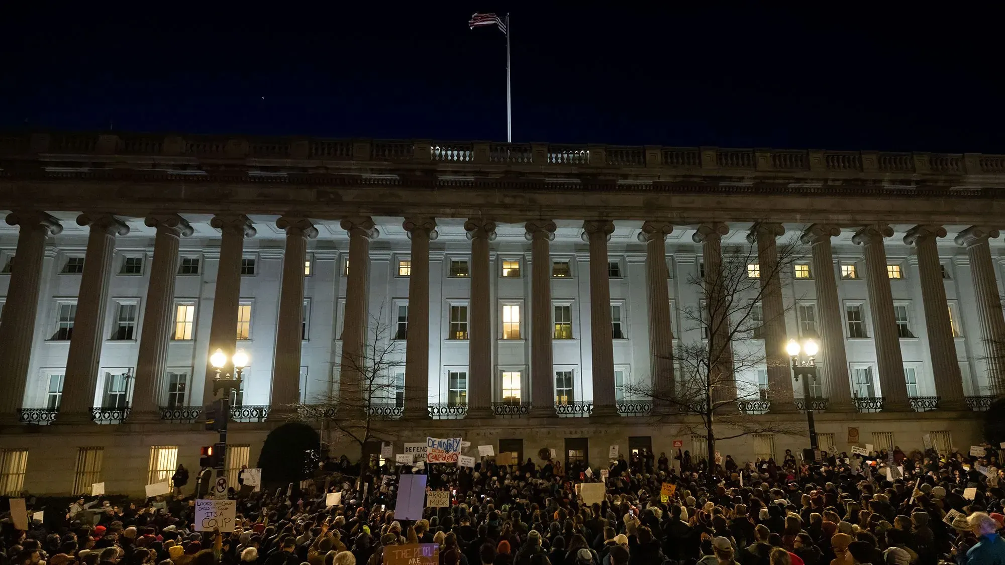 مظاهرة حاشدة أمام مبنى حكومي مضاء ليلاً، حيث يجتمع المتظاهرون حاملين لافتات تعبر عن احتجاجاتهم على سياسات إدارة ترامب.