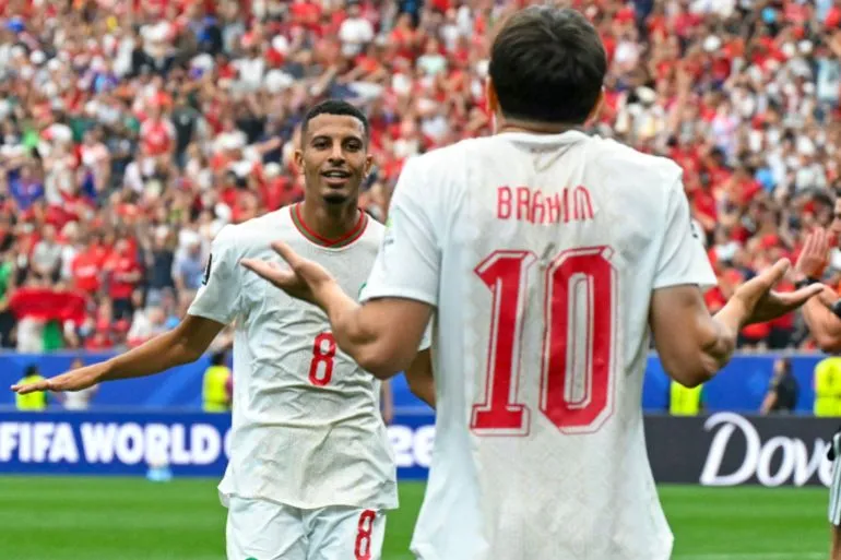 لاعبا المنتخب المغربي يحتفلان بهدف في مباراة كأس العالم وسط حماس الجماهير، مع التركيز على تفوق المغرب في البطولة.
