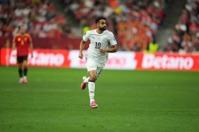 لاعب كرة قدم يرتدي قميصًا أبيض يحمل الرقم 10، يسير في الملعب خلال مباراة، مع جمهور خلفه وأضواء ساطعة، في أجواء كأس العالم.
