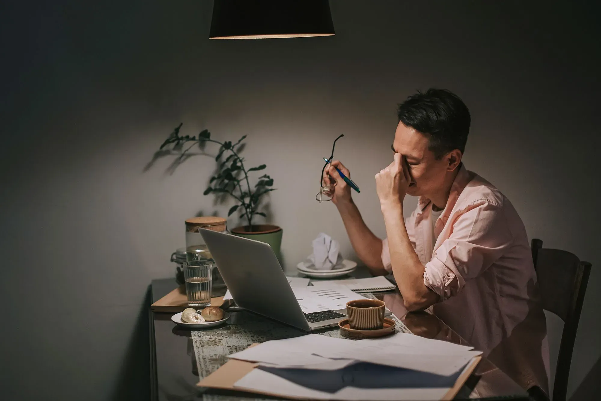 رجل يجلس على طاولة في غرفة مضاءة بشكل خافت، يبدو عليه القلق والإجهاد المالي، محاط بأوراق ومعدات مكتبية.