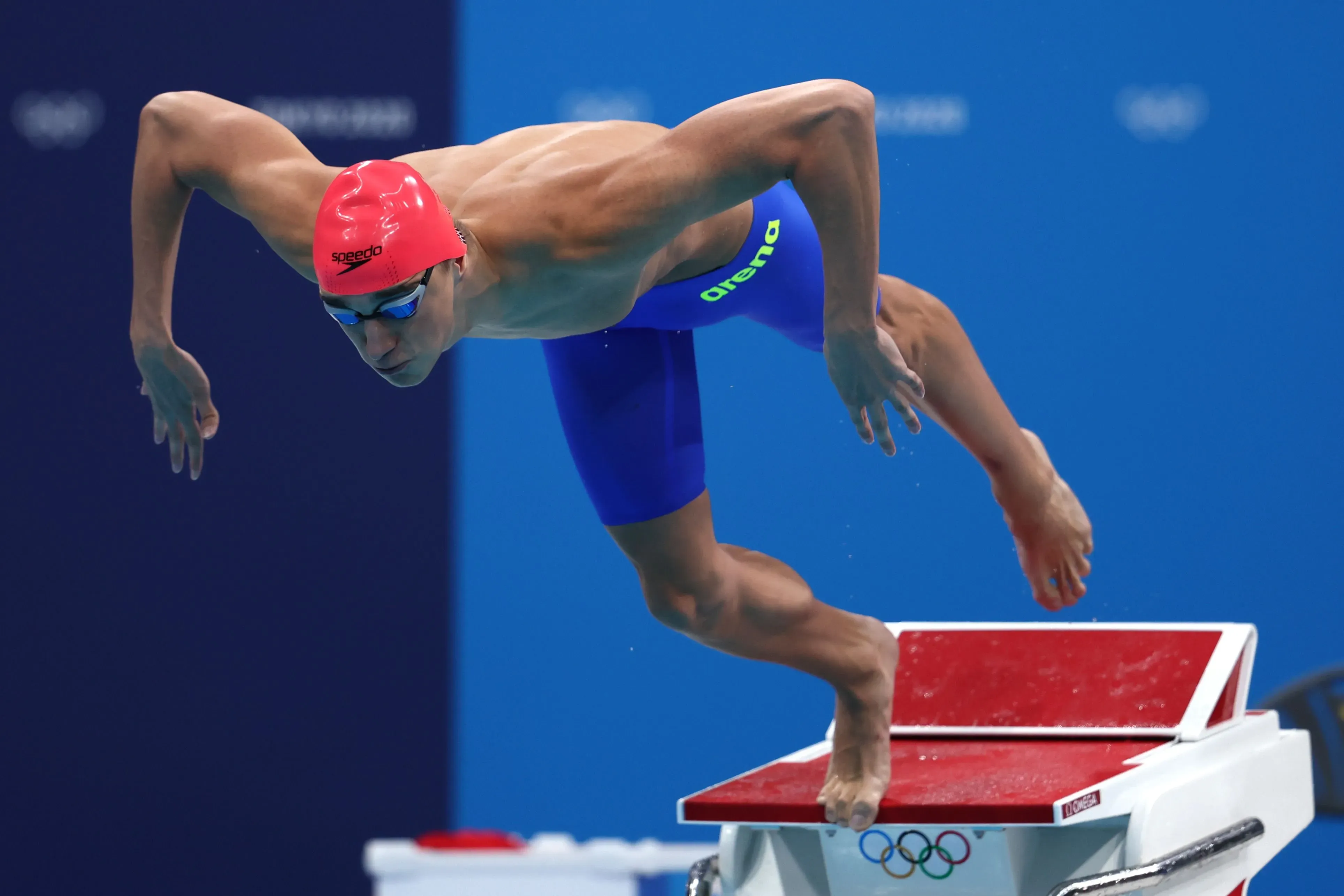 سباح أوكراني في وضع الانطلاق، يرتدي قبعة حمراء ونظارات سباحة، يستعد للقفز في سباق 50 مترًا حرة، مع خلفية زرقاء.