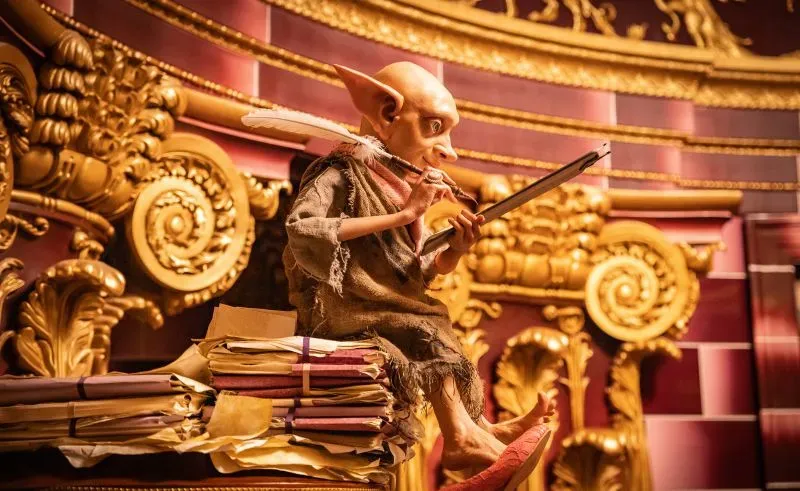 شخصية خيالية تشبه الجني تجلس وسط أكوام من الملفات في بيئة مزخرفة، تعكس أجواء وزارة السحر من عالم هاري بوتر.