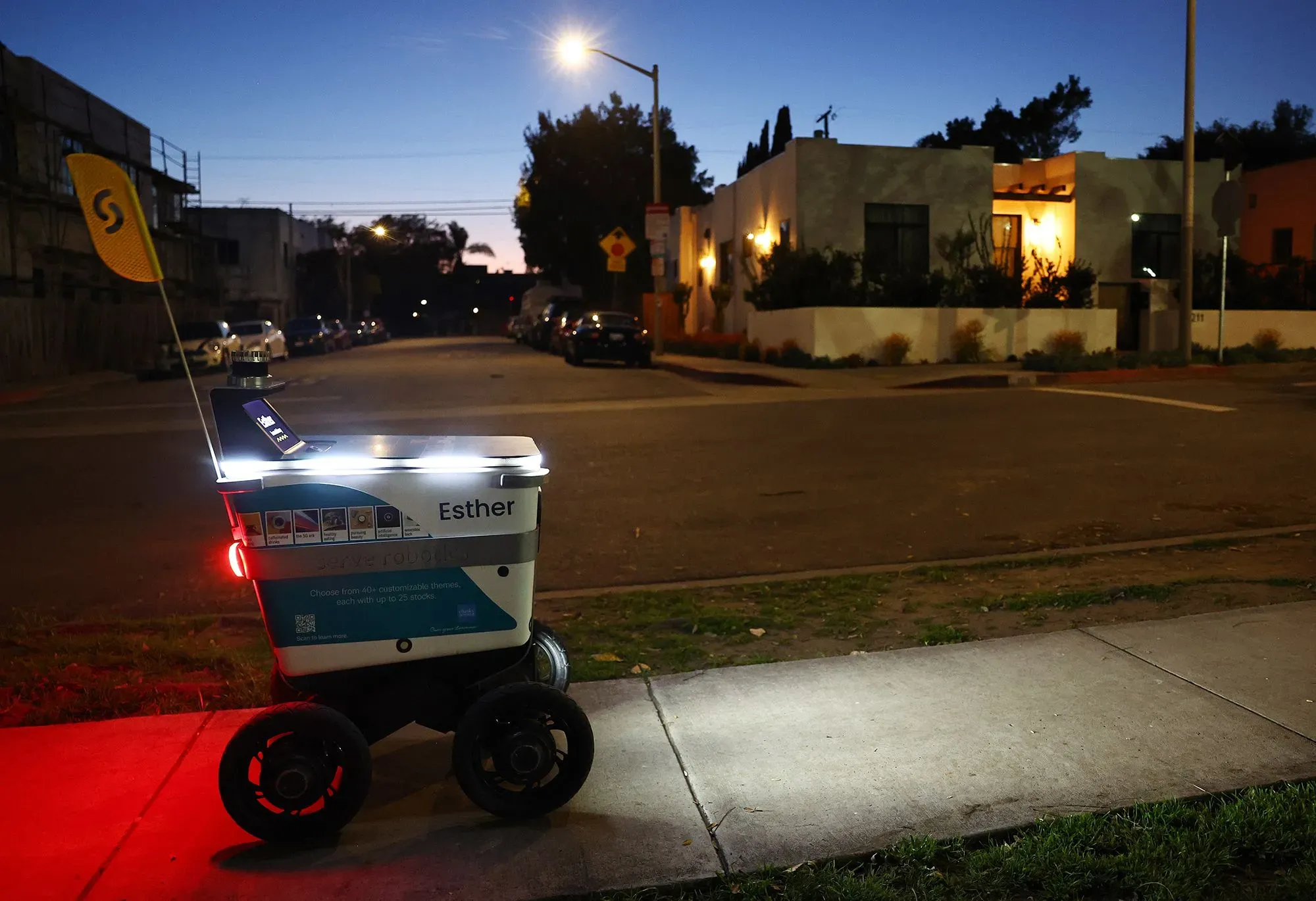 روبوت توصيل الطعام "كورتني" يضيء بمصابيح أمامية في حي سكني بأتلانتا، مع وجود منازل خلفه، يظهر كجزء من الحياة اليومية.