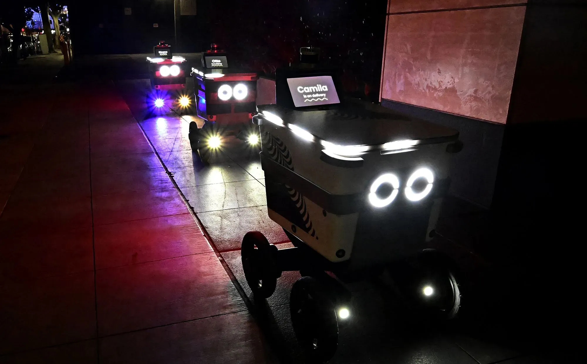 روبوتات توصيل الطعام ذات العجلات الأربعة، مضاءة بأضواء ساطعة، متوقفة في الشارع، تعكس تطور التكنولوجيا في خدمات التوصيل.