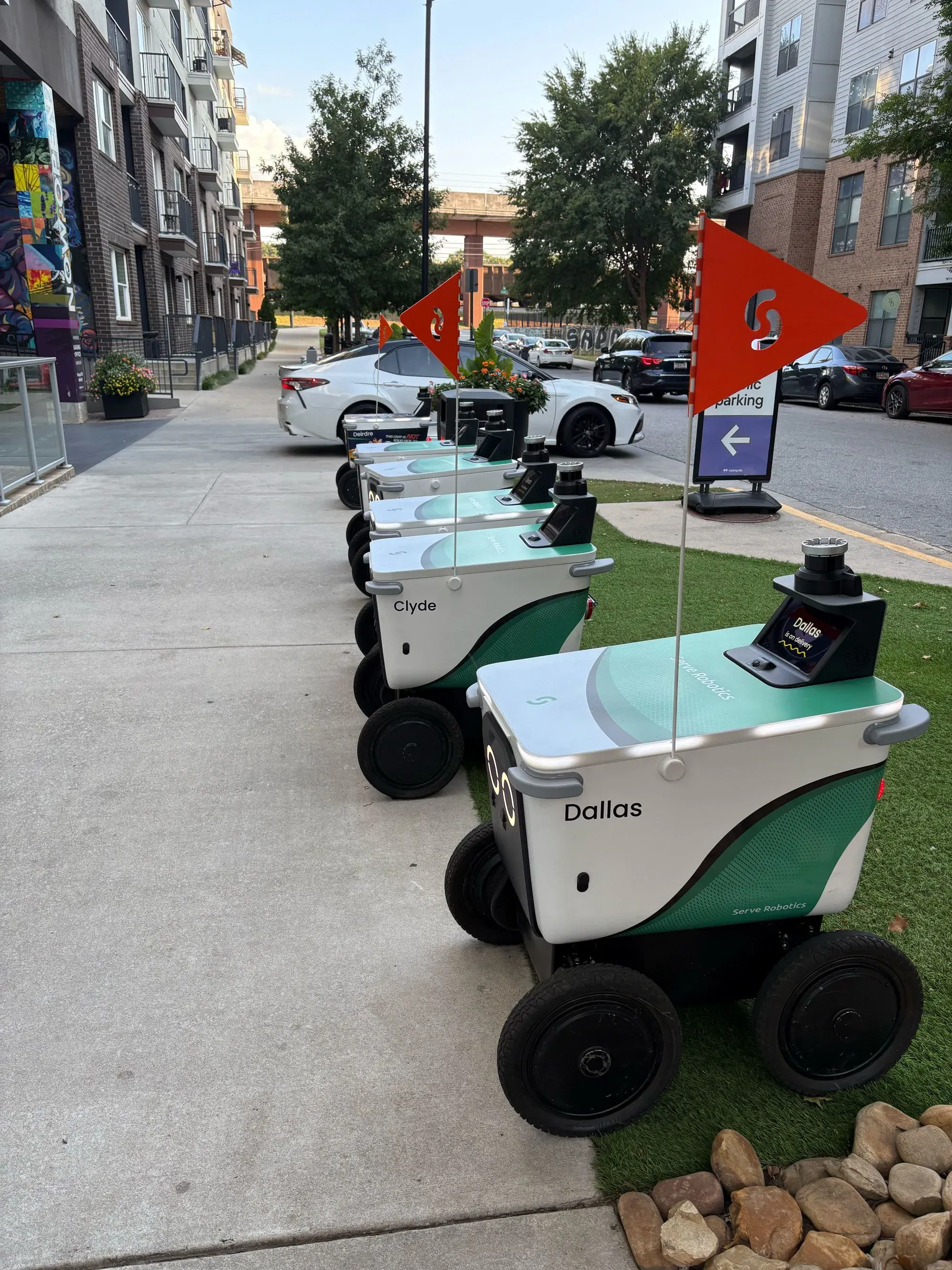 روبوتات توصيل الطعام من شركة Serve Robotics، مصطفة على الرصيف في أتلانتا، تحمل أسماء مثل "دالاس" و"كلايد"، مع أعلام حمراء.