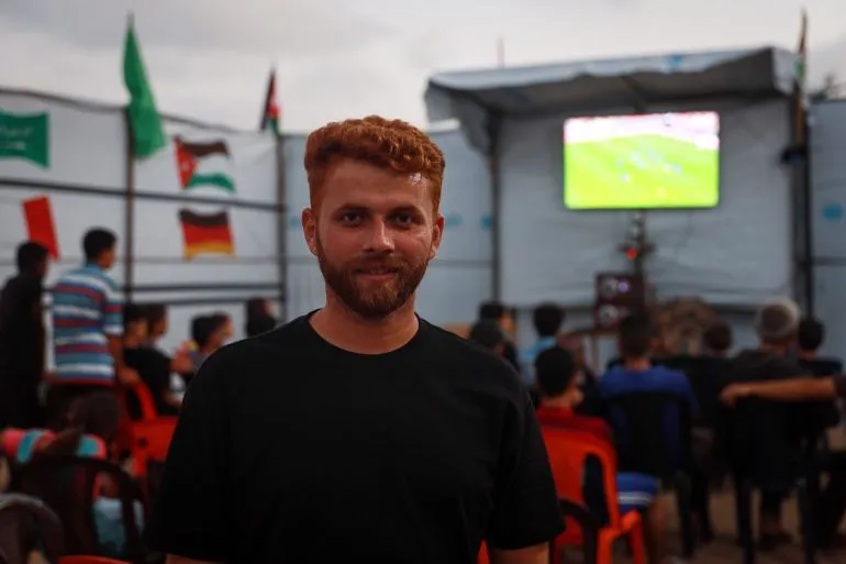 شاب مبتسم يقف أمام خيمته في استاد اليرموك بغزة، بينما تُعرض مباراة كأس العالم على شاشة خلفه، وسط أجواء من التوتر والتهجير.