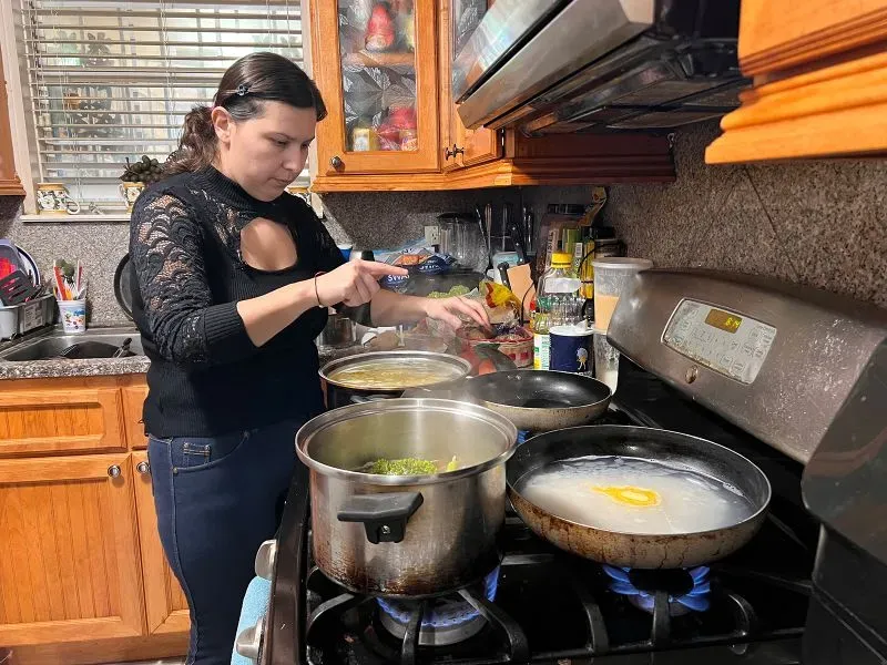 امرأة تعد الطعام في مطبخها، حيث تظهر أواني الطهي المختلفة على الموقد، مع التركيز على الجهود لتلبية احتياجات أسرتها الغذائية.