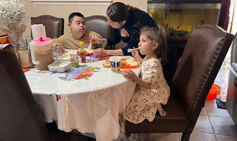 امرأة تساعد طفلين على تناول الطعام حول طاولة مغطاة بمفرش مزخرف، مع وجود أطباق وأكواب بجوارهم، تعكس تحديات الحياة اليومية.