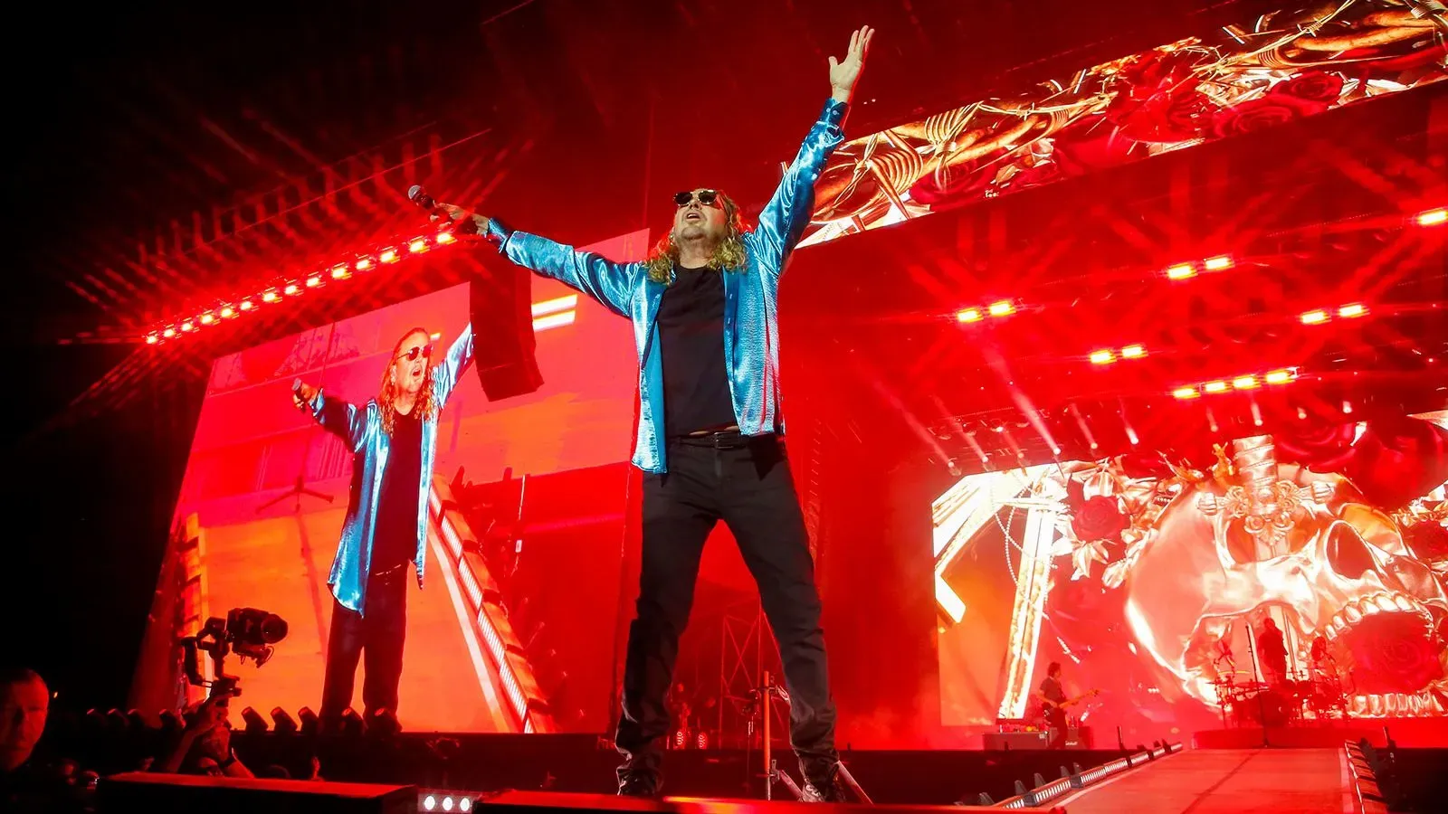 مغني فرقة Maná يؤدي على المسرح خلال عرض حماسي، مع أضواء حمراء ساطعة وشاشة خلفية تعرض رسومات ملونة.