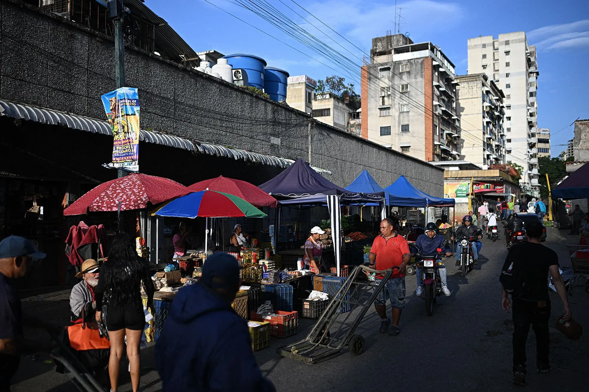 سوق مزدحم في فنزويلا، حيث يتجول الناس بين أكشاك الطعام تحت مظلات ملونة، مع خلفية لمباني سكنية. تعكس الصورة الحياة اليومية والتحديات الاقتصادية.