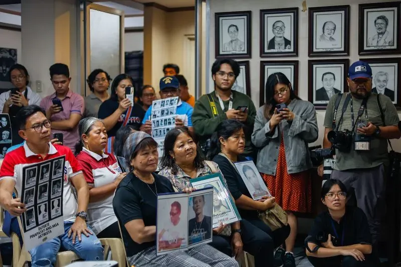 مجموعة من الأشخاص يجلسون في قاعة، يحملون صور ضحايا حرب المخدرات في الفلبين، مع وجود لوحات على الجدران تعكس معاناة العائلات.