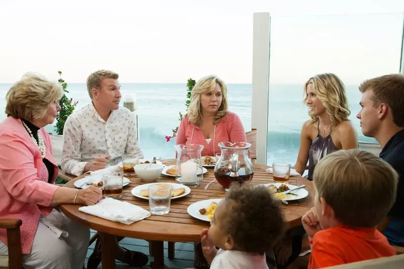 عائلة كريسلي تتناول الطعام معًا على شرفة تطل على البحر، مع أجواء عائلية دافئة وتفاعل بينهم، يعكس حياتهم الثرية.