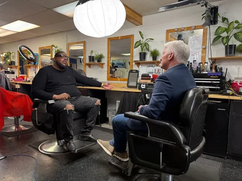 مقابلة في صالون حلاقة، حيث يتحدث رجل ذو نظارات مع شخص آخر. تبرز خلفية الصالون مع مرايا ونباتات.