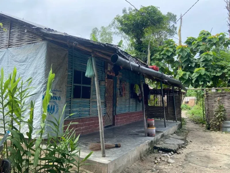 منزل بسيط في مخيم للاجئين، محاط بالنباتات، يمثل الظروف المعيشية الصعبة للاجئين من بوتان بعد ترحيلهم.