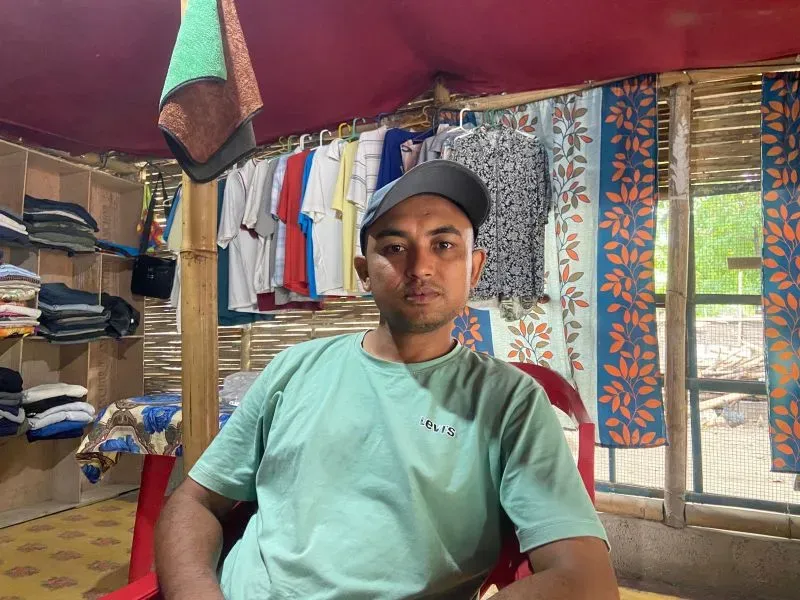 شاب يجلس في مخيم للاجئين، محاطًا بالملابس المعلقة، يعبر عن صعوبة الحياة بلا وثائق بعد ترحيله من الولايات المتحدة إلى بوتان.
