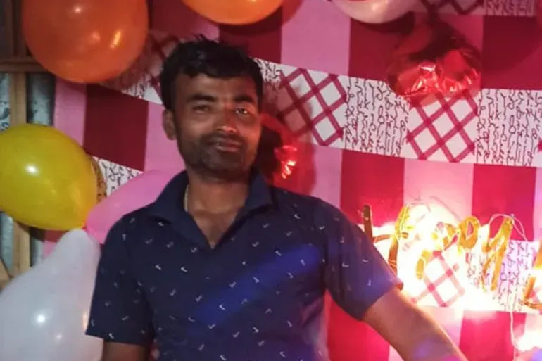 صورة لرجل يقف أمام خلفية مزينة بالبالونات، مع عبارة "عيد ميلاد سعيد" مضاءة، تعكس أجواء احتفالية في سياق معاناة الهجرة.