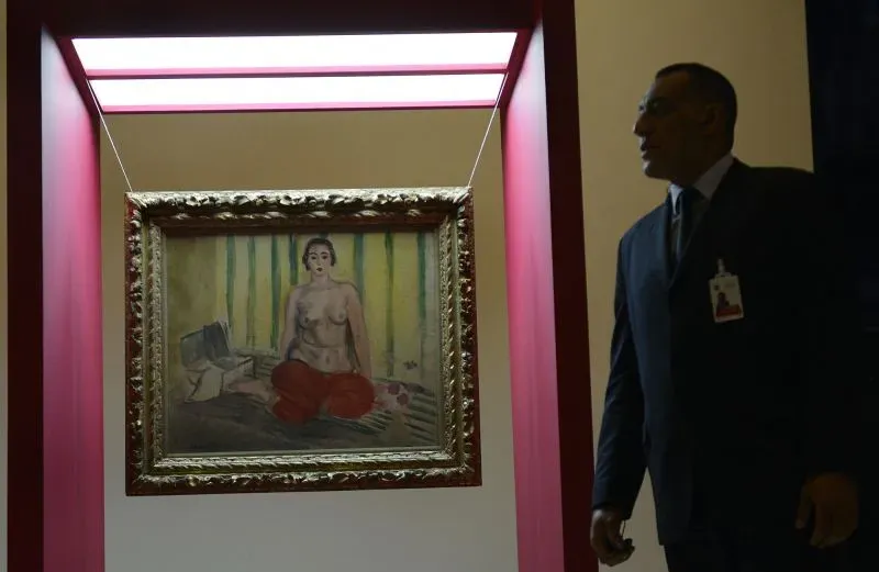 لوحة فنية ملونة تُظهر امرأة جالسة في وضعية تأملية، محاطة بإطار ذهبي، بينما يقف حارس أمن بجانبها.