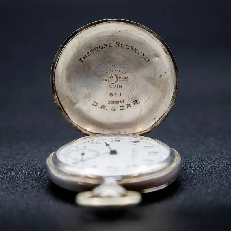 ساعة جيب قديمة مفتوحة تُظهر وجه الساعة الأبيض مع عقارب تشير إلى الوقت، وداخل الغطاء نقش يحمل اسم "ثيودور روزفلت" ورقم 911.