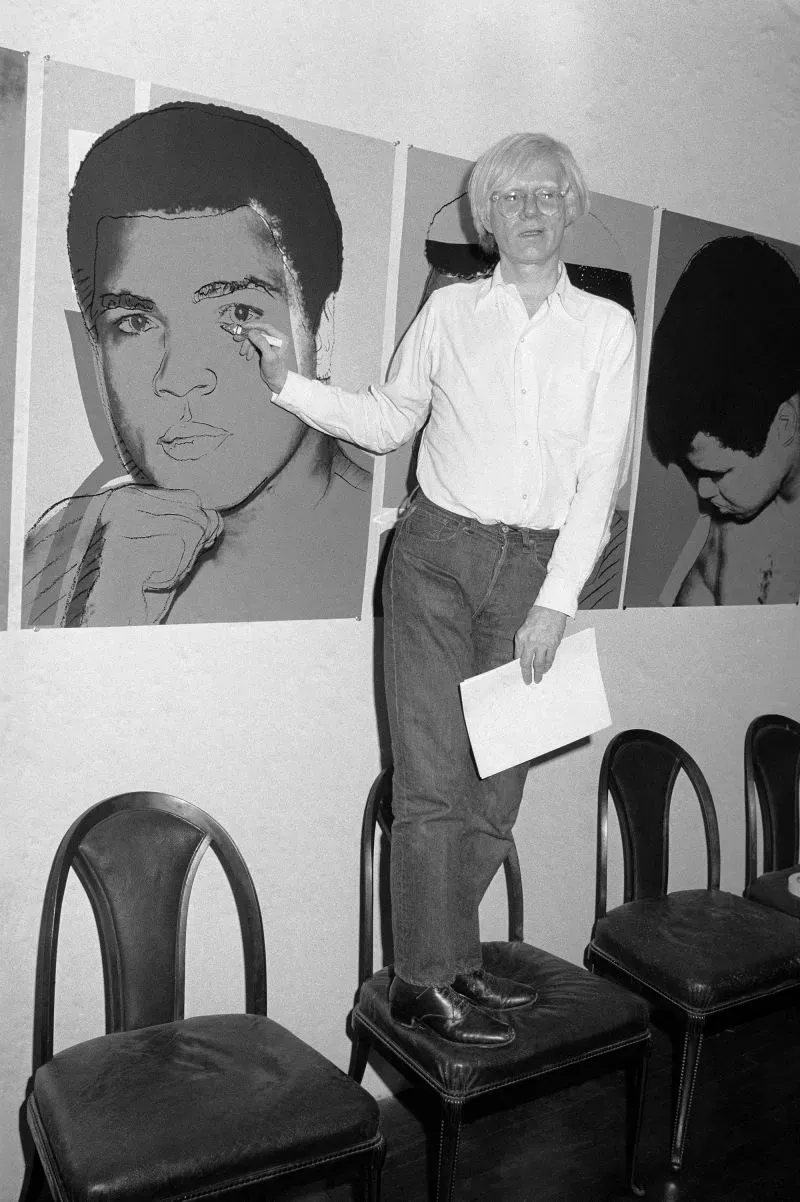 فنان يقف على كراسي، يعمل على لوحة شهيرة لمحمد علي، مع صورته خلفه، مما يعكس تأثيره في عالم الفن والثقافة.