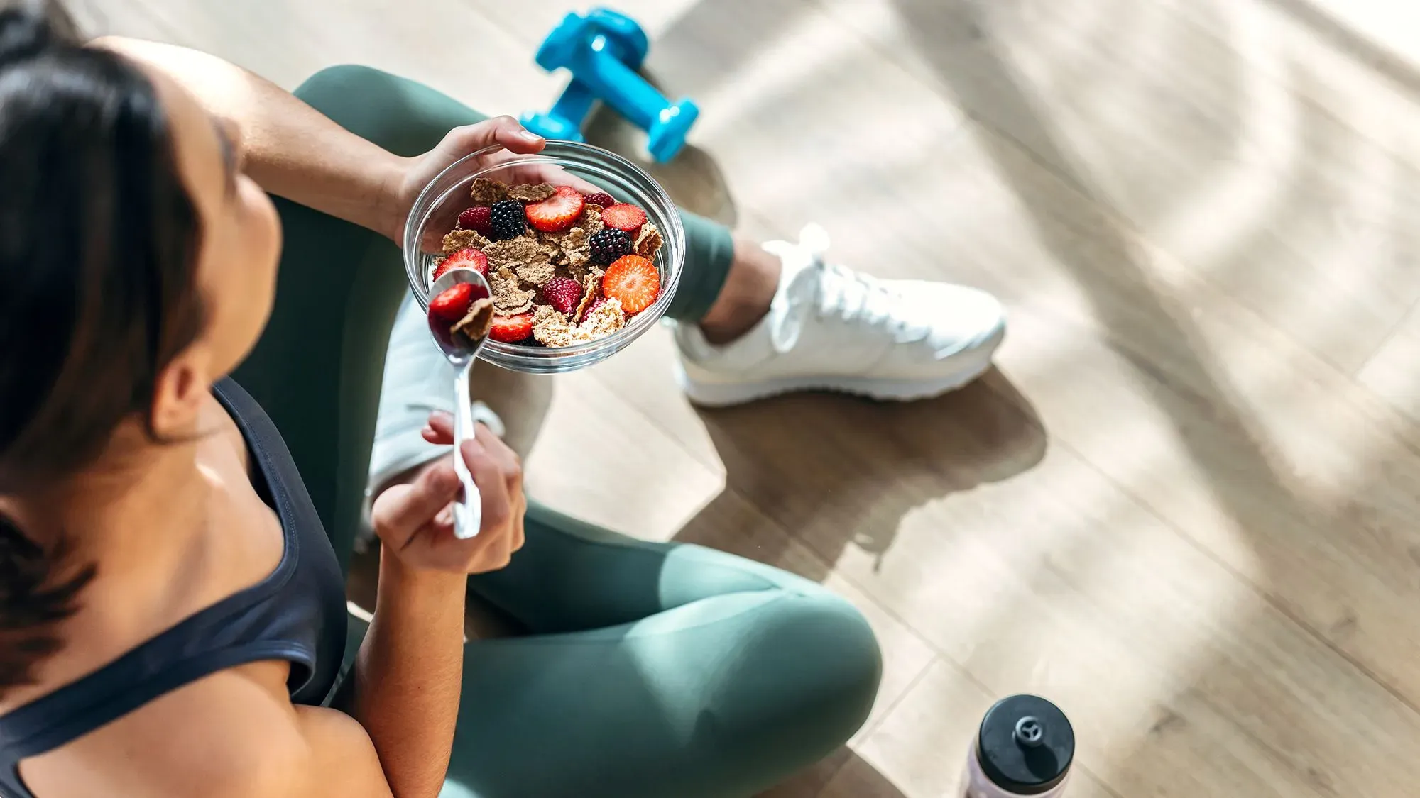 امرأة تجلس على الأرض وتتناول وعاء يحتوي على الشوفان مع الفواكه، محاطة بأدوات رياضية، مما يشير إلى أهمية تكوين عادات صحية.