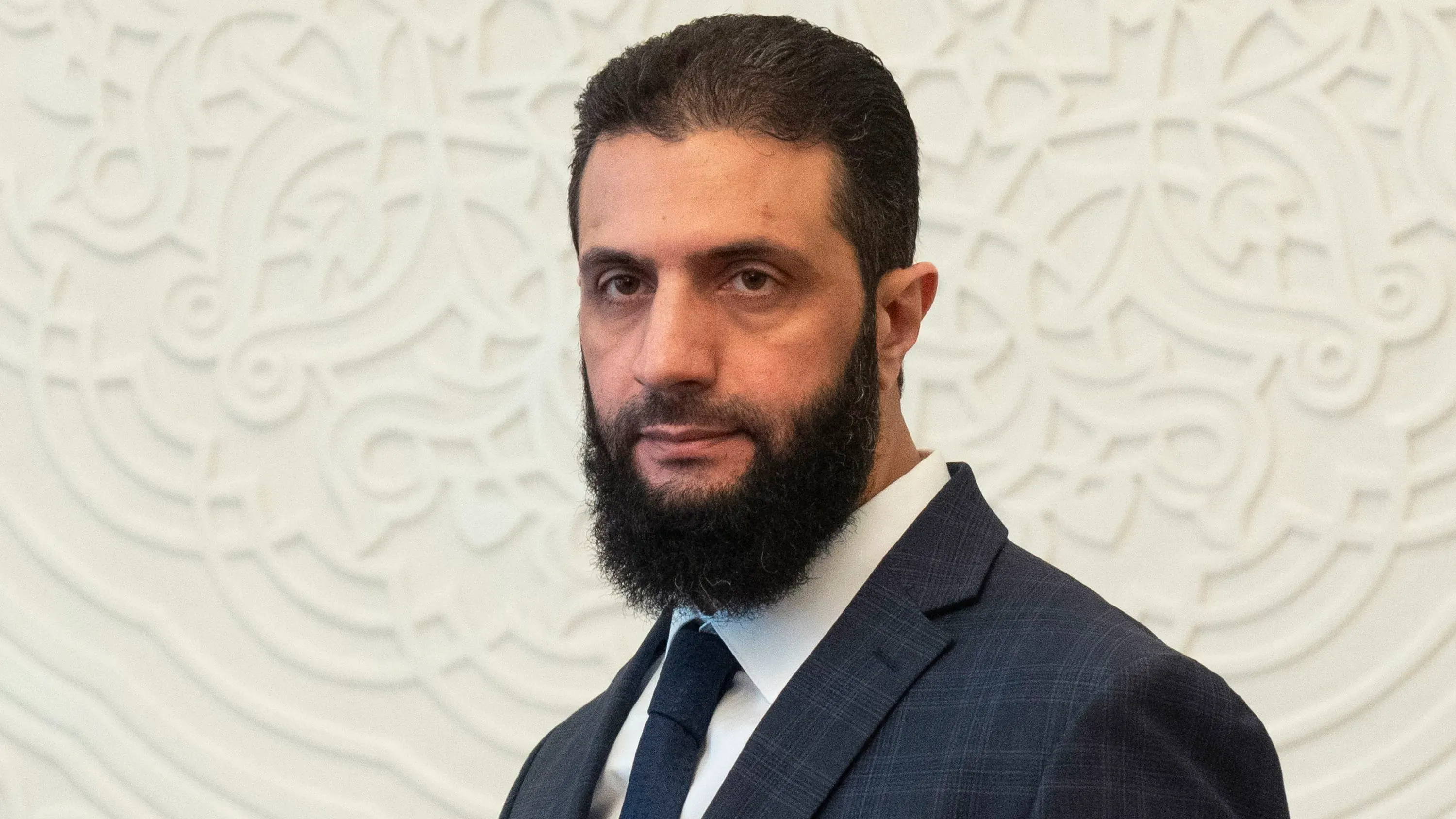 أحمد الشرع، المعروف سابقًا بأبو محمد الجولاني، في مؤتمر صحفي يعلن توليه رئاسة سوريا خلال فترة انتقالية، مع خلفية زخرفية.