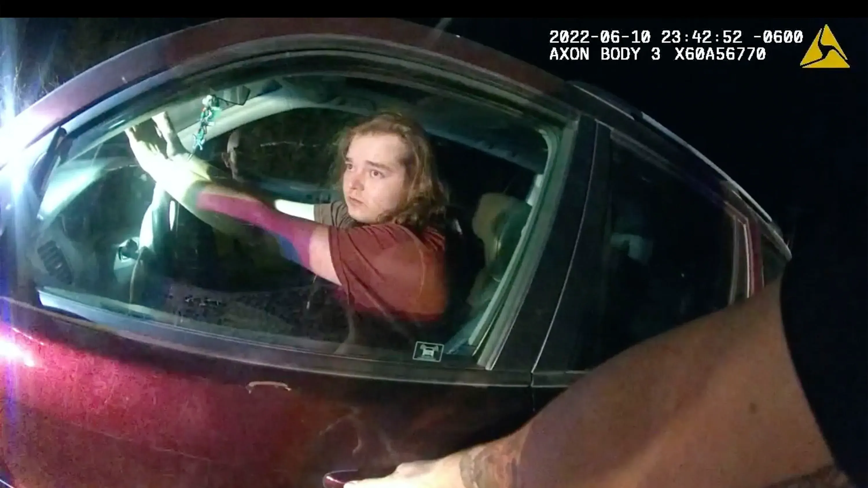 شخص يظهر داخل سيارة حمراء، يبدو عليه القلق، بينما تتواجد أضواء الشرطة في الخلفية، في سياق حادثة إطلاق نار تتعلق بالصحة العقلية.