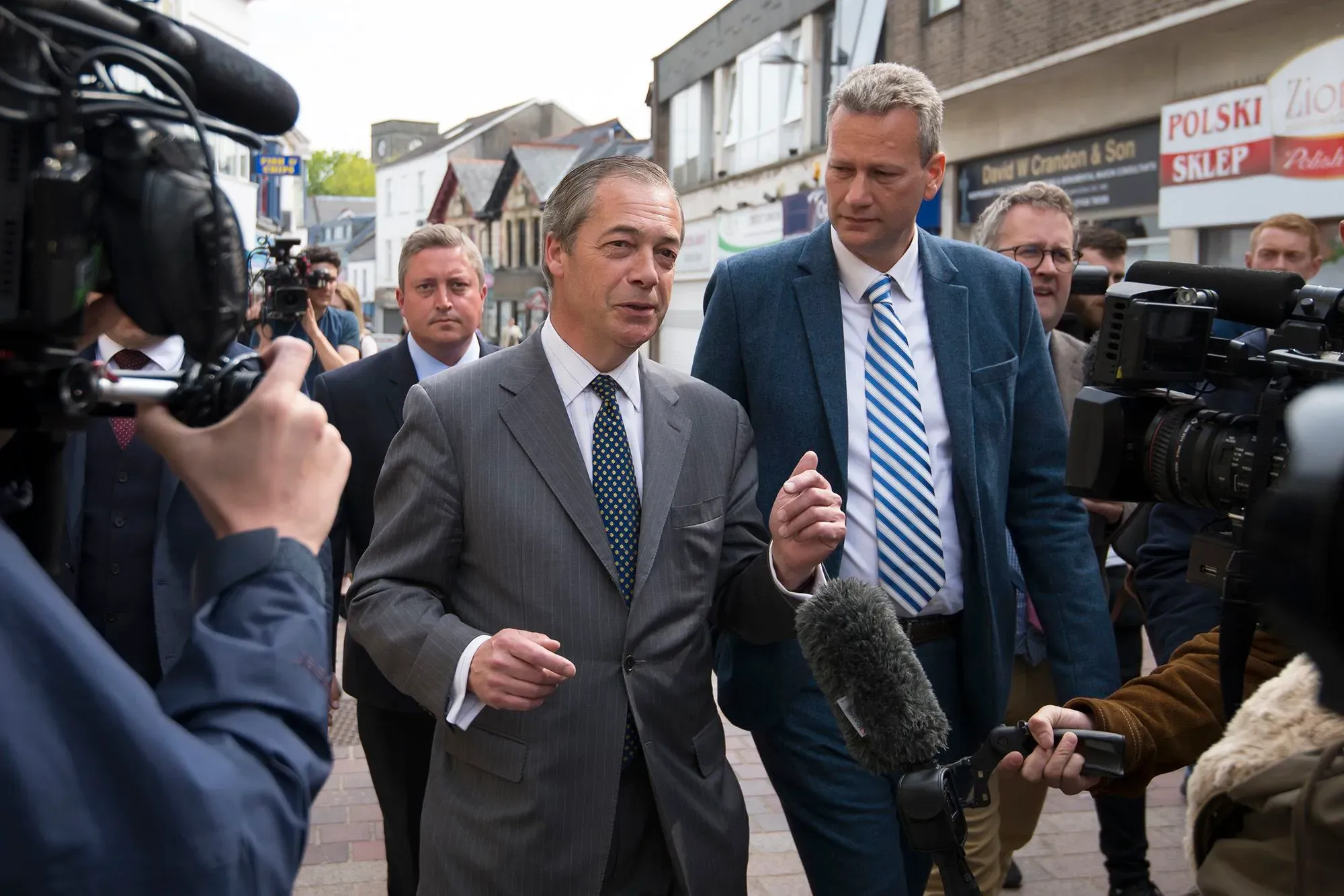 نايجل فاراج، زعيم حزب الإصلاح البريطاني، يتحدث إلى الصحفيين في الشارع، محاطًا بمجموعة من الأشخاص والكاميرات، في سياق فضيحة سياسية.