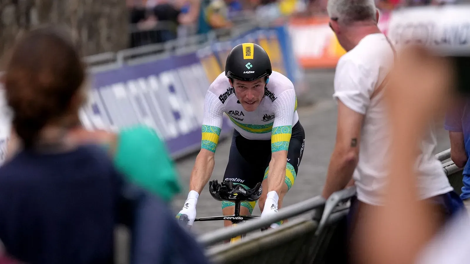 الدراج الأوليمبي روهان دينيس يتسابق في حدث رياضي، مرتديًا زي فريقه، مع حشود من المشجعين في الخلفية.