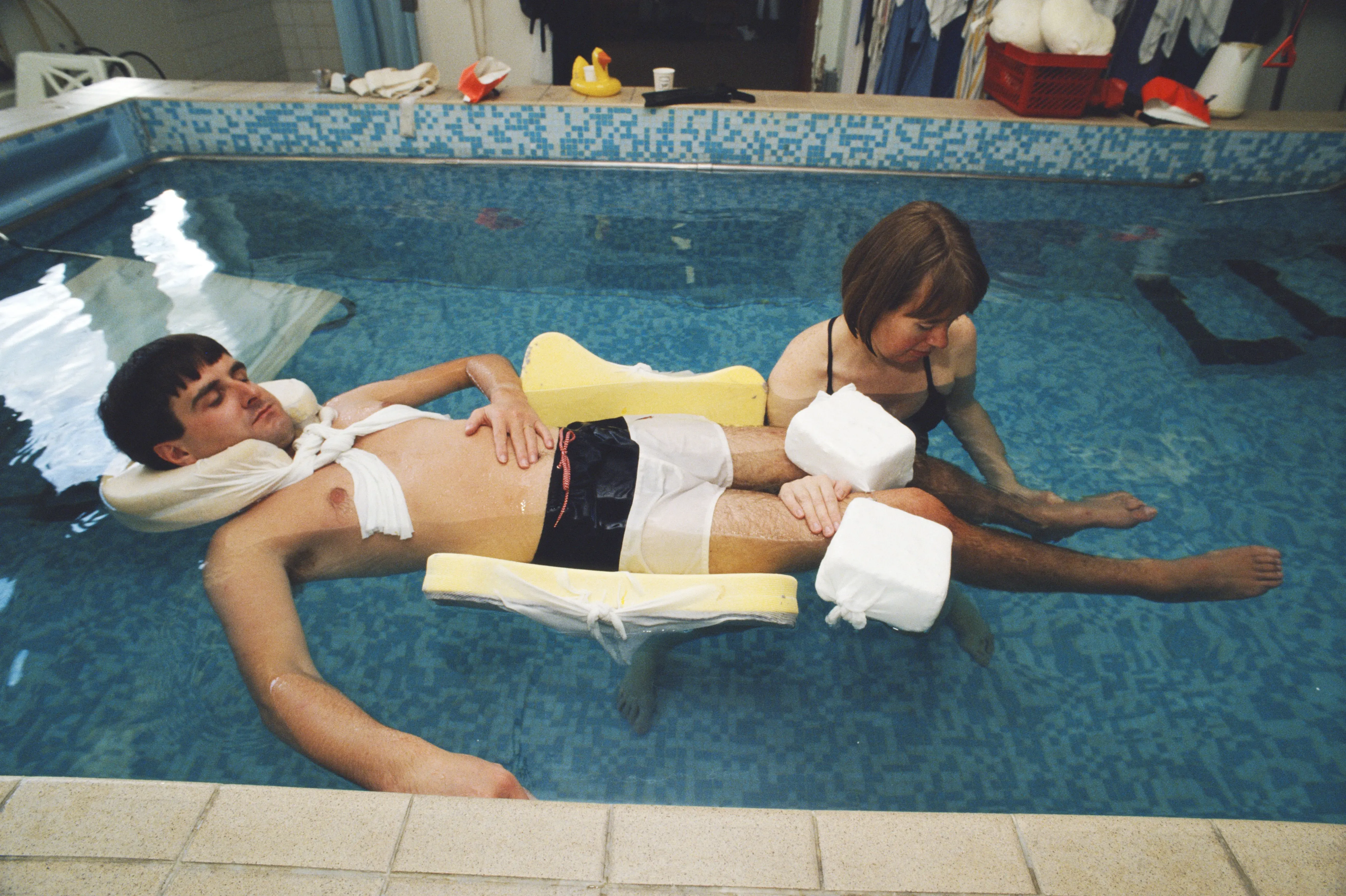 شاب مستلقٍ في بركة سباحة، يظهر عليه آثار إصابة، بينما تساعده امرأة في عملية إعادة التأهيل.