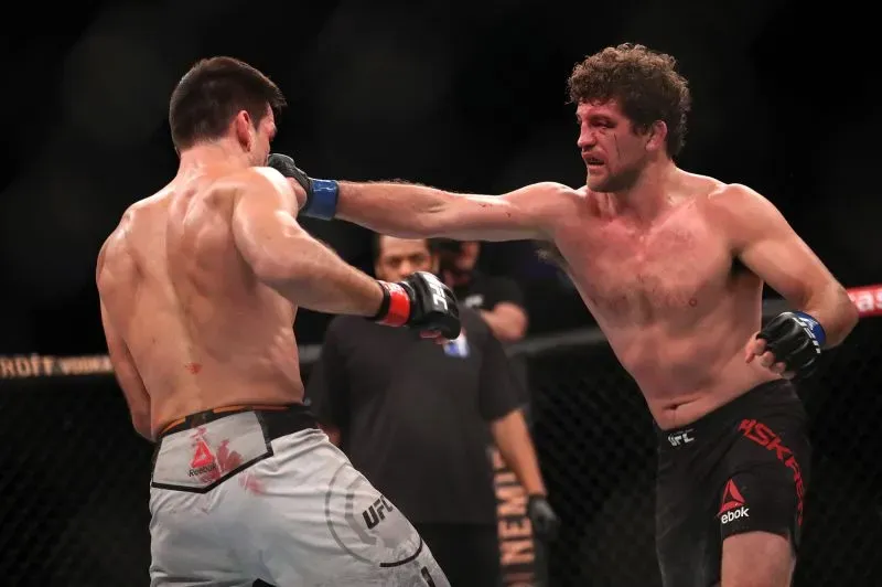 مقاتل الفنون القتالية المختلطة بن أسكرين يوجه ضربة لمنافسه خلال نزال في UFC، مع وجود حكم في الخلفية.