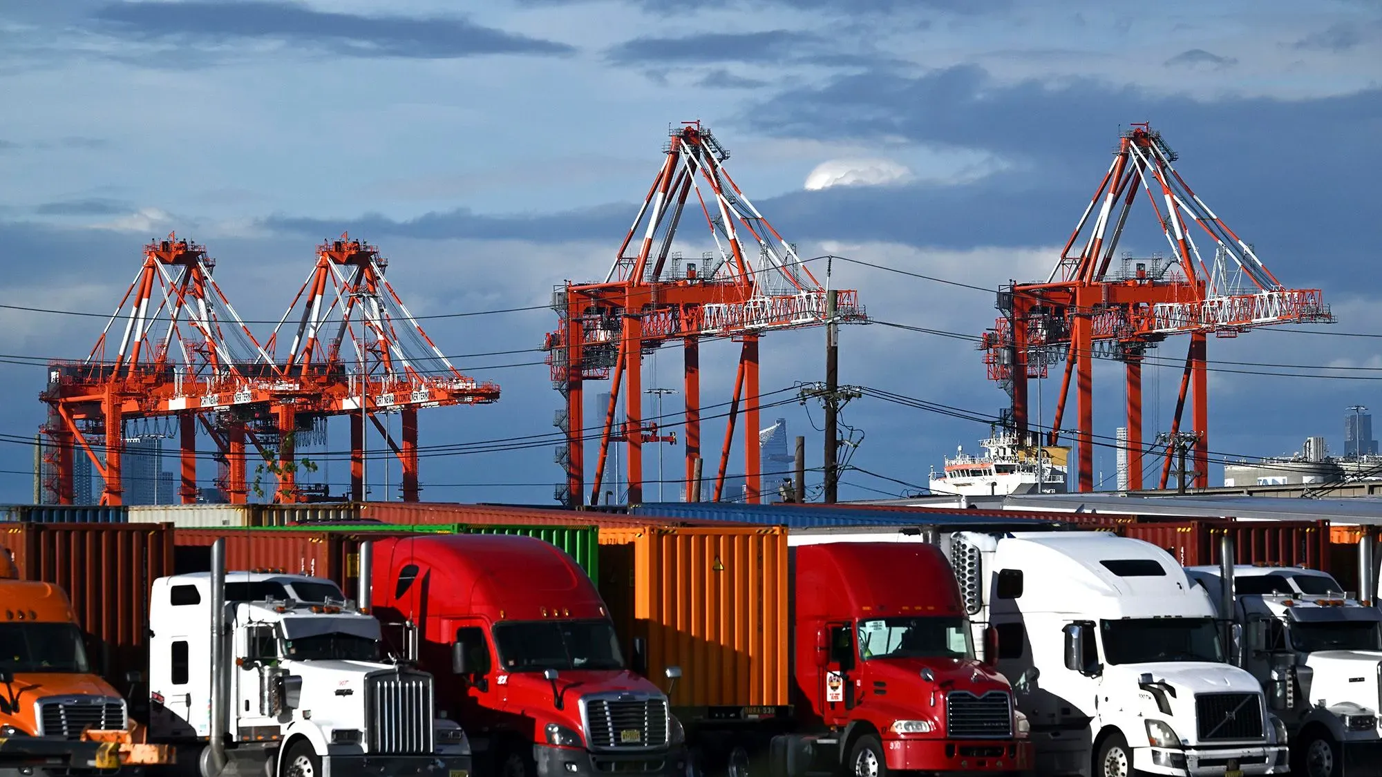 صورة لميناء شحن مزدحم، تظهر شاحنات نقل حمراء وبيضاء وقوائم رافعات برتقالية، تعكس تأثير الرسوم الجمركية على التجارة الأمريكية.