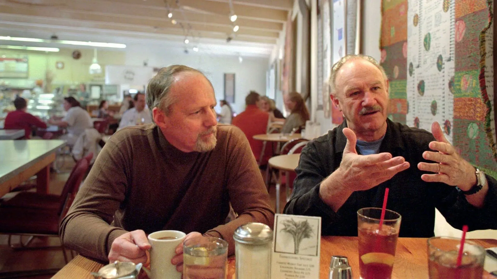 لقاء ودّي في مطعم، حيث يتحدث رجلان معًا، مع وجود زبائن آخرين في الخلفية، مما يعكس لحظة من التواصل الاجتماعي.