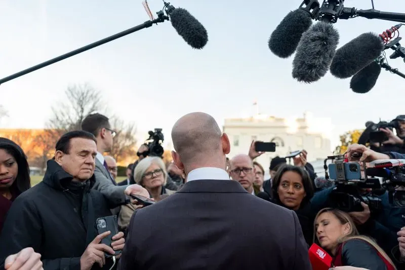 ستيفن ميلر، مستشار ترامب، يتحدث إلى حشد من الصحفيين أمام البيت الأبيض، محاطًا بميكروفونات وكاميرات، بينما تسلط الأضواء عليه.