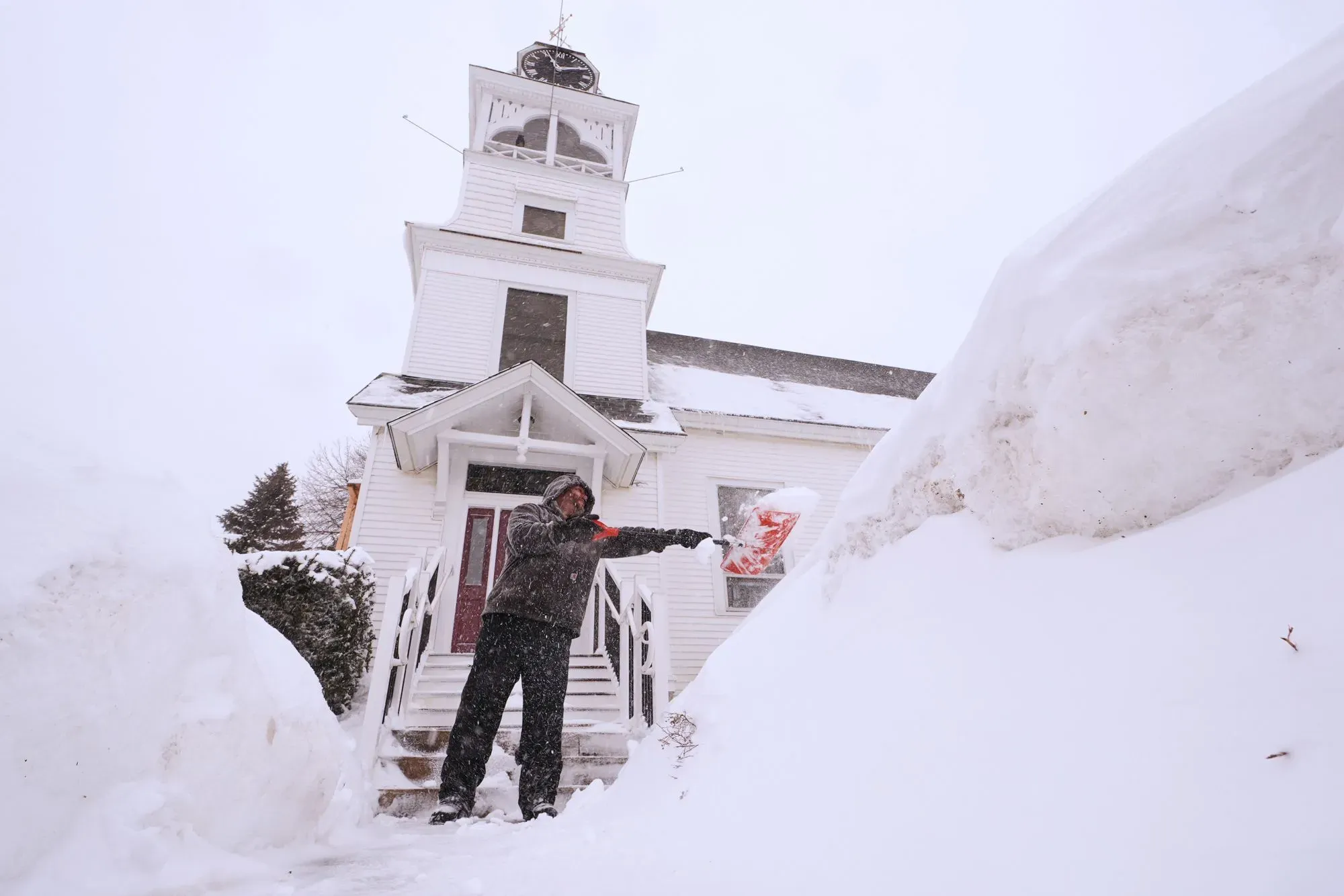 رجل يستخدم مجرفة لإزالة الثلوج من أمام مبنى خلال عاصفة شتوية قوية، مع تراكم الثلوج حوله بشكل كبير.
