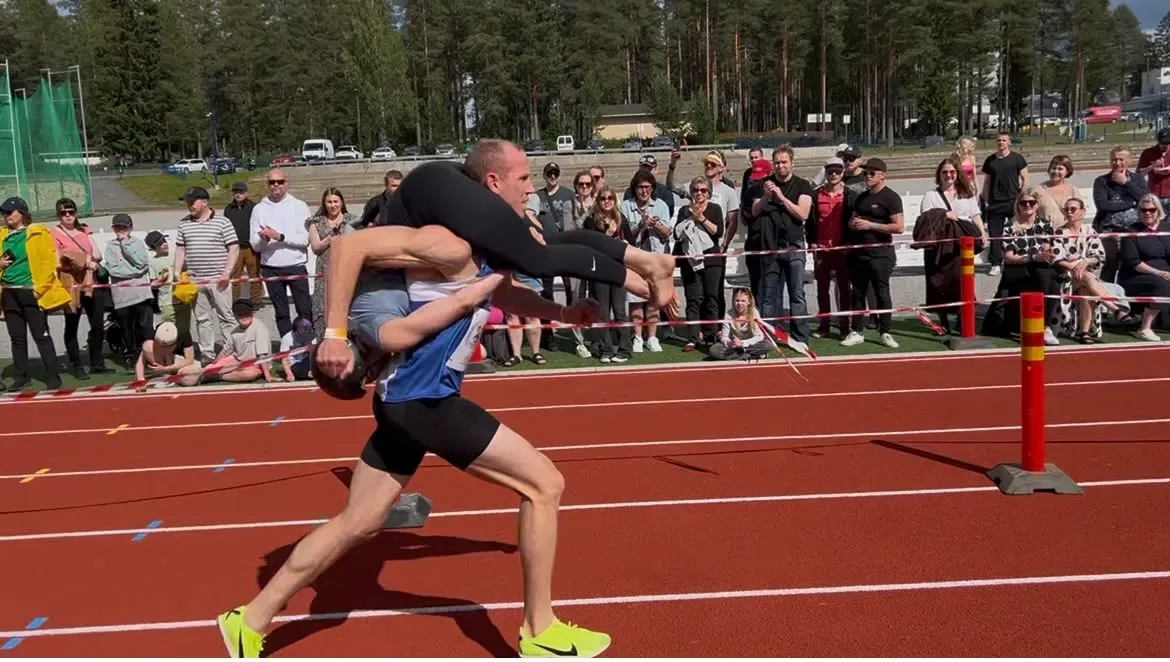 زوج يحمل زوجته على كتفيه أثناء سباق حمل الزوجات، مع جمهور يشاهد في الخلفية. تعكس الصورة أجواء المنافسة والمرح في هذا الحدث الفريد.