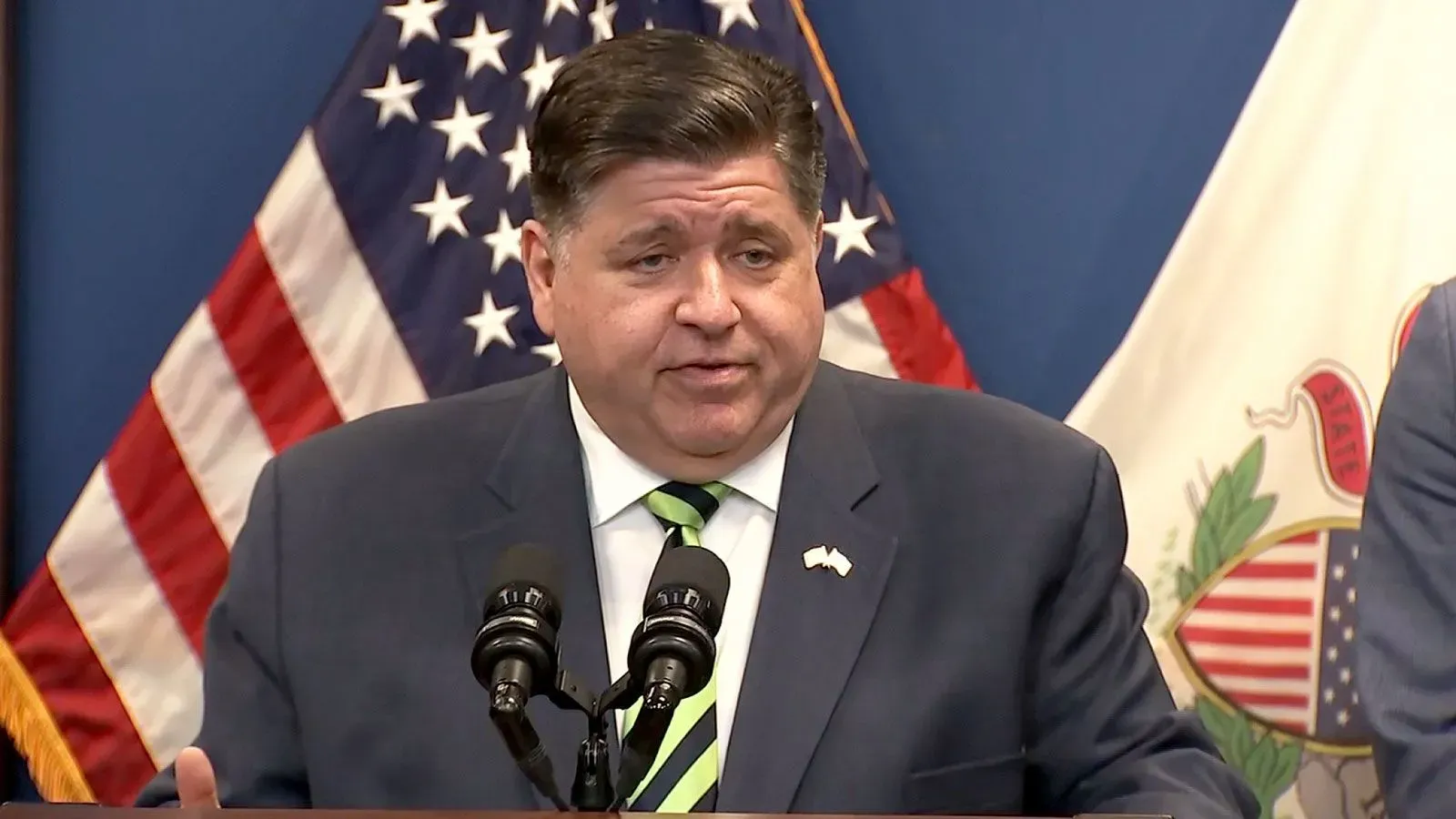 حاكم إلينوي جي بي بريتزكر يتحدث خلال مؤتمر صحفي، مع العلم الأمريكي خلفه، معبرًا عن معارضته لتجميد المساعدات الفيدرالية.