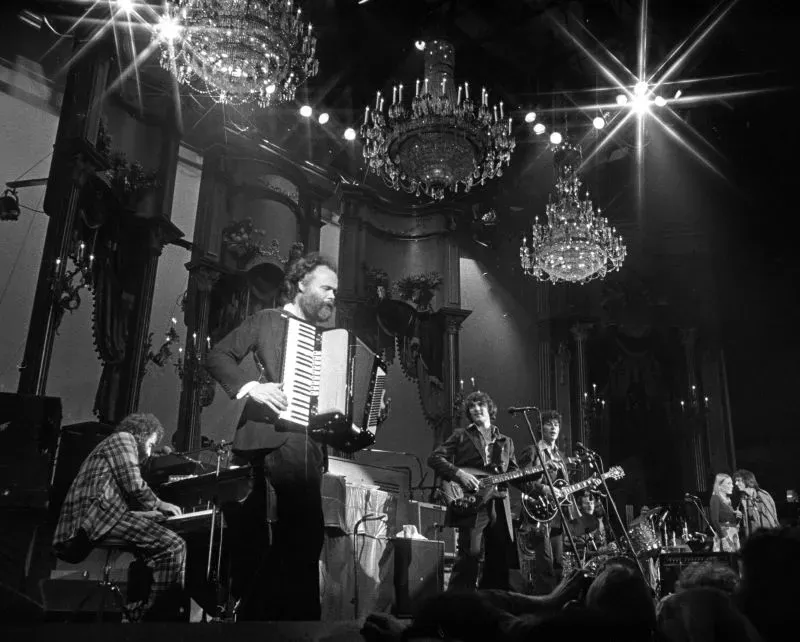 غارث هدسون، عازف الكيبورد في فرقة ذا باند، يؤدي على المسرح مع أعضاء آخرين، محاطًا بأضواء لامعة وثريات.