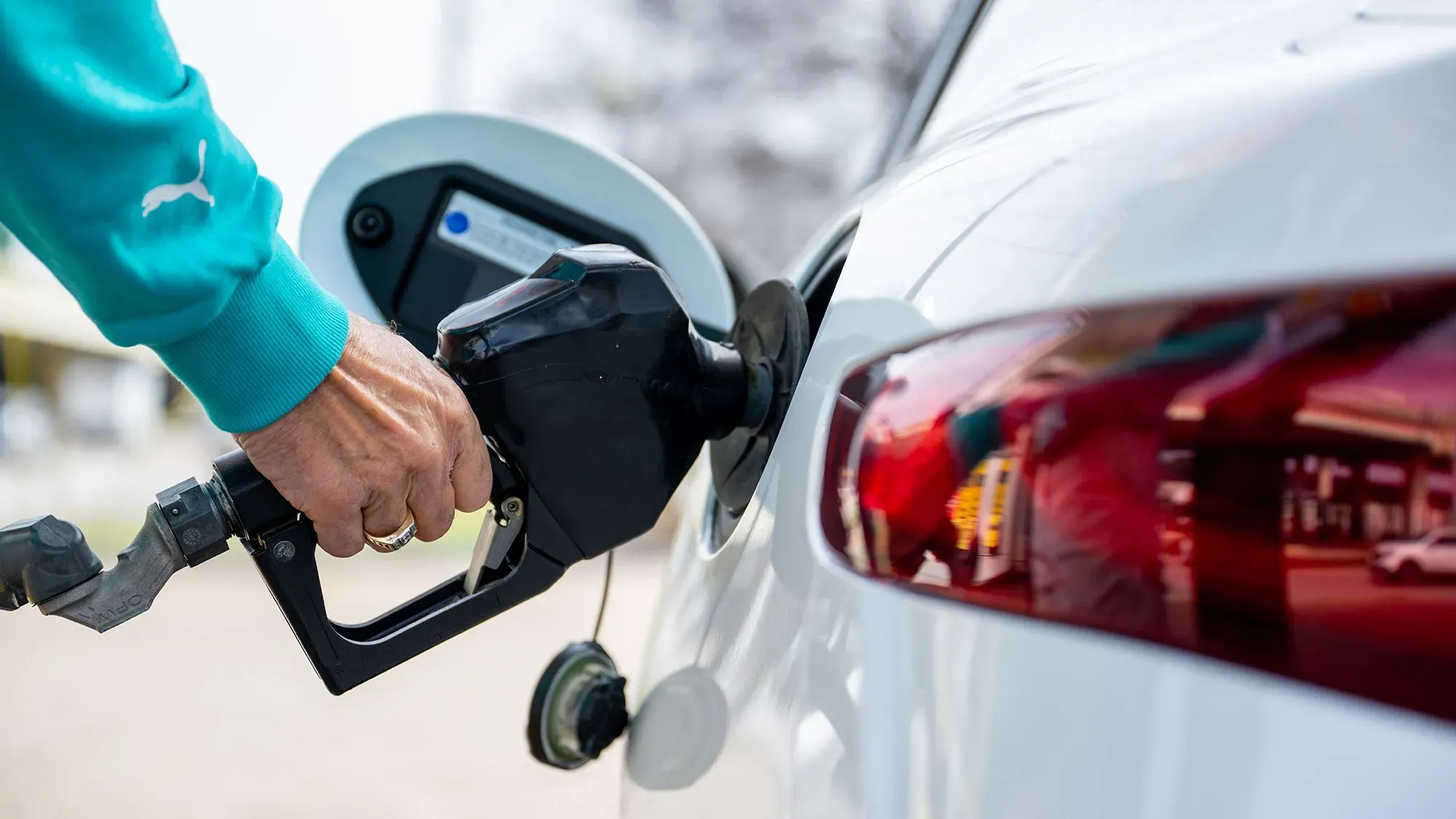 شخص يملأ خزان وقود سيارة بيضاء في محطة وقود، مع التركيز على انخفاض أسعار البنزين وتأثيره على الاقتصاد الأمريكي.