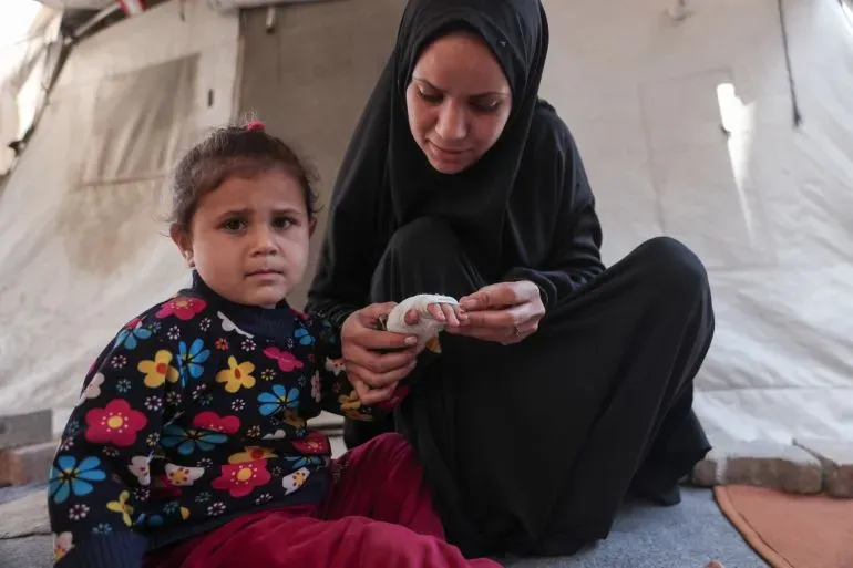 سماح الدبلة تعالج جرح ابنتها ميّاسين في خيمتهما المؤقتة بجوار جبل من الأنقاض، وسط مخاوف من الجرذان التي تهاجمهم.