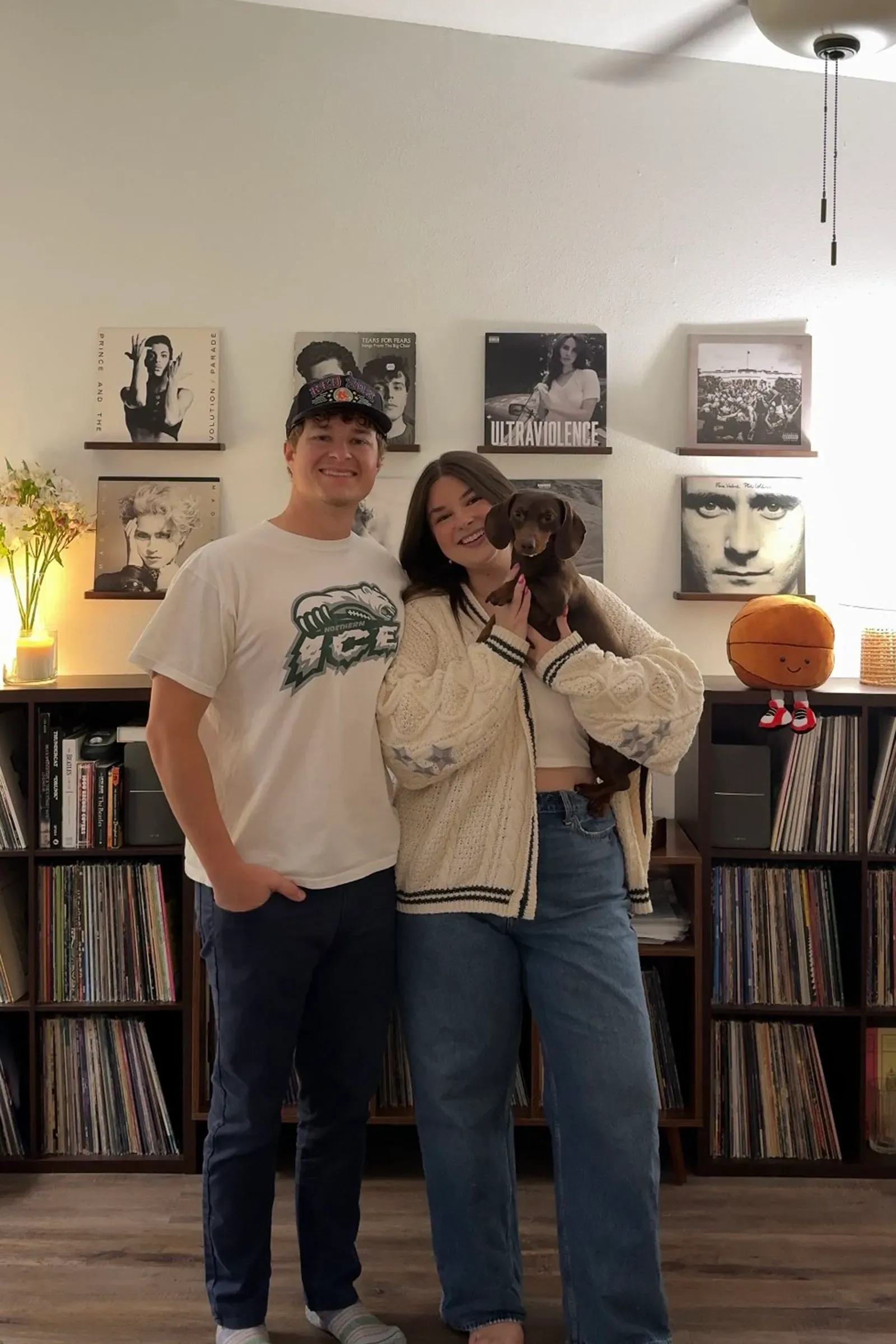 زوجان شابان يقفان مع كلبهما أمام رف مليء بأسطوانات الفينيل، مع إضاءة دافئة تضفي جواً مريحاً على المكان.