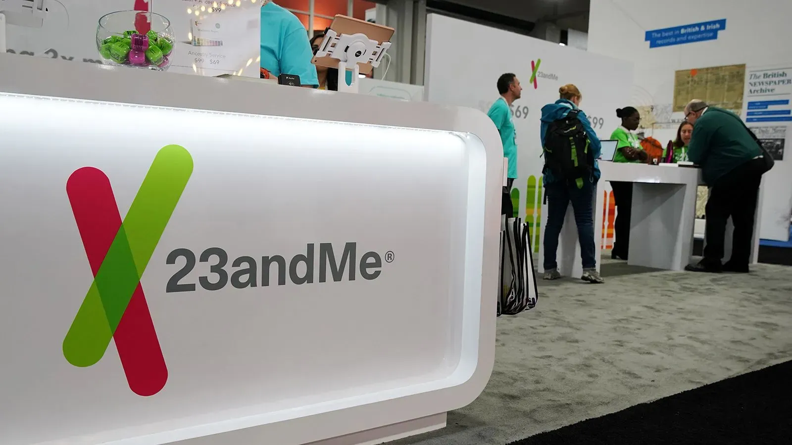 شعار شركة 23andMe يظهر في معرض، مع موظفين يتفاعلون مع الزوار، في سياق إعلان الشركة عن الإفلاس وإعادة الهيكلة.